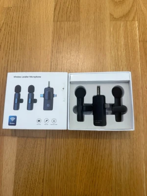 Trådlös Lavalier Mikrofon - Säljer en svart trådlös lavalier mikrofon med två små mikrofoner och en mottagare. Perfekt för inspelning av video, podd eller streaming. Mikrofonerna är kompakta och har rund form, levereras i originalförpackning med tydliga ikoner och instruktioner.