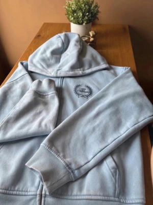 Ljusblå hoodie med dragkedja - Säljer en ljusblå hoodie med dragkedja och huva. På bröstet finns ett broderat motiv med texten 'Not Ceremony' inramat av en krans. Hoodien har ribbade muddar vid ärmar och nederkant. Perfekt för en chill och avslappnad stil.