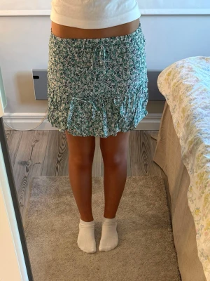 Grön blommig kortkjol från Zara - Grön kortkjol från Zara med vitt blommönster och volangkant. Kjolen har snörning i midjan och shorts under kjolen.