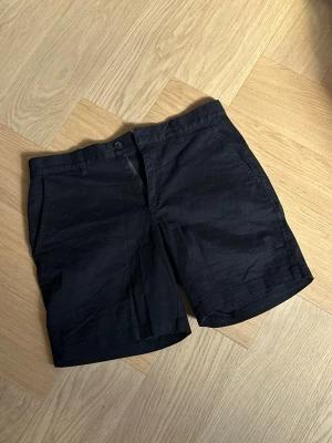 Svarta klassiska shorts i bomull från H&M - Säljer ett par svarta shorts i bomull med normal passform. De har knapp och dragkedja framtill, bälteshällor och fickor både fram och bak. Perfekta för sommaren och enkla att matcha med olika stilar.