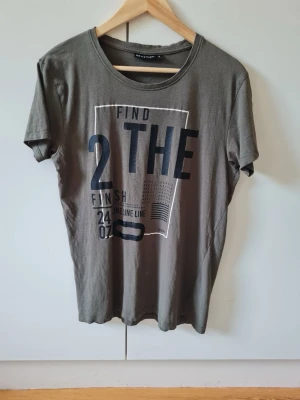 Grå t-shirt med tryck från Reaction strl S - Säljer en grå t-shirt från Reaction med grafiskt tryck på framsidan i svart och vitt. T-shirten har rund halsringning och korta ärmar. Material 100% bomull. Storlek S.
