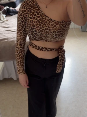 Leopardmönstrad oneshoulder croppad topp - Säljer en leopardmönstrad croppad topp med oneshoulder och lång ärm. Toppen har en snygg omlott-detalj med knyt runt midjan som ger en unik look. 