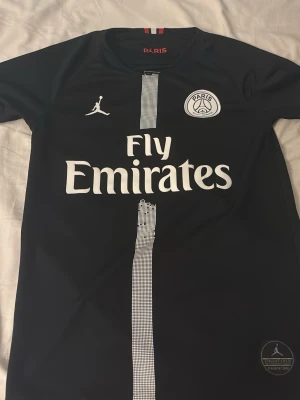 PSG tröja - Snygg svart PSG matchtröja i samarbete med Jordan. Tröjan har vita detaljer, PSG-logga och Fly Emirates-tryck på bröstet samt en vertikal rand i mitten. Tillverkad i lätt och ventilerande material, perfekt för fotboll eller träning.