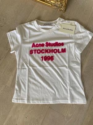Vit Acne Studios t-shirt med tryck S - Vit t-shirt från Acne Studios med rosa texttryck på bröstet där det står 'Acne Studios STOCKHOLM 1996'. Klassisk rund hals och korta ärmar. Tillverkad i mjuk bomull för en skön och avslappnad look.
