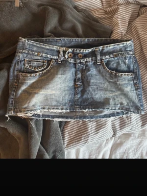 Snygg  jeanskjol - Säljer en kort blå jeanskjol med slitna detaljer och rå, fransig kant nedtill. Kjolen har dubbla knappar i midjan, fickor fram och bak samt nitar längs fickkanterna för en extra edgy look. Perfekt för dig som gillar en avslappnad och trendig stil.