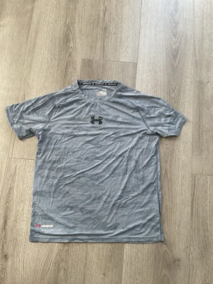 Grå träningströja från Under Armour - Snygg grå kortärmad träningströja från Under Armour med diskret kamouflagemönster. Tillverkad i ett lätt och snabbtorkande funktionsmaterial som passar perfekt till gymmet eller löprundan. Logga på bröstet och liten logga nere vid fållen.