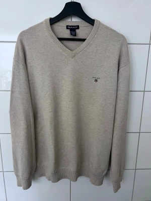 Beige stickad V-ringad tröja Gant - Beige tröja från Gant med V-ringning och broderad logga på bröstet. Tröjan har ribbade muddar vid ärmslut och nederkant för en snygg passform. Perfekt för dig som gillar klassisk och stilren design.
