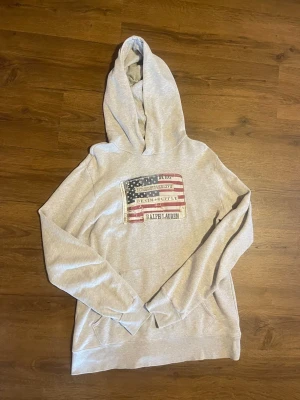 Grå Ralph lauren hoodie - Vintage grå Ralph lauren x denim supply hoodie med snyggt tryck. Den tidigare ägaren har klippt resprbandet vid midjan, så priset är definitivt diskuterbart. Trots dessa defekter så har tröjan en bra passform.