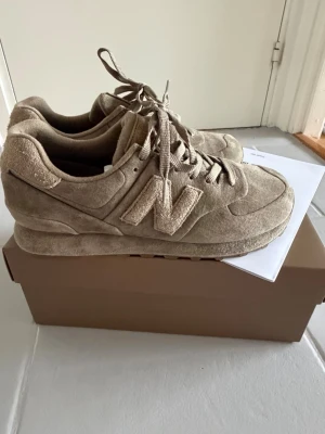 Beige New Balance x Stone Island 574 sneakers - Snygga beige New Balance 574 sneakers i mocka med klassisk N-logga på sidan. Skorna har en låg siluett, matchande snören och en chunky sula för extra komfort. Perfekta för dig som gillar stilrena och neutrala färger.