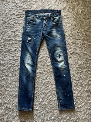 Blå slitna jeans från Dsquared2 - Säljer ett par blåa jeans från Dsquared2 med slitningar, hål och färgstänk för en riktigt edgy look. Jeansen har smal passform, klassiska fem fickor och detaljerade sömmar. Perfekta för dig som vill sticka ut med din stil.
