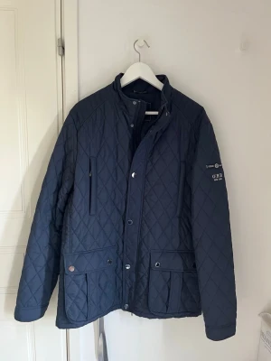 Mörkblå quiltad jacka med fickor - Snygg mörkblå quiltad jacka med hög krage och tryckknappar. Jackan har två stora framfickor med lock, en bröstficka med dragkedja och detaljer på ärmen. Perfekt för dig som gillar klassisk men modern stil.
