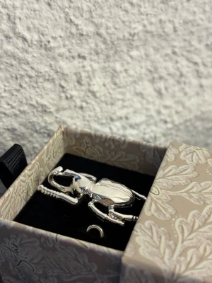 Silverfärgad ring IOAKU - Unik justerbar ring i form av en skalbagge i blank silverfärg. Broschen har detaljerade ben och kropp, och levereras i en ask. Perfekt accessoar för dig som vill sticka ut med något annorlunda och coolt smycke. Från IOAKU