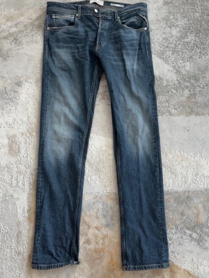  Replay grover - Säljer ett par klassiska blå jeans från Replay med raka ben och fem fickor. Jeansen har snygga slitningar och kontrastsömmar samt en liten röd Replay-detalj vid fickan. Tillverkade i denim med en autentisk tvättad look. Grover modell