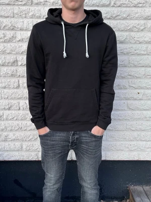 Svart hoodie med vita snören - Snygg svart hoodie med klassisk känguruficka och vita dragsnören i huvan. Perfekt för en avslappnad streetstyle-look. Hoodien har ribbade muddar vid ärmar och nederkant samt en mjuk insida för extra komfort.