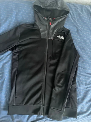 Svart hoodie från The North Face - Snygg svart hoodie från The North Face med dragkedja hela vägen, huva och logga på bröstet. Materialet är en mix av mjukt tyg och glansigare paneler upptill. Praktiska fickor och detaljer i rött vid dragkedjan ger extra stil.
