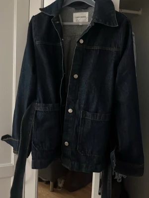 Mörkblå jeansjacka med bälte - Snygg mörkblå jeansjacka med klassisk krage, stora framfickor och knäppning framtill. Jackan har ett matchande bälte i midjan och långa ärmar med knappdetaljer vid ärmsluten. Perfekt för en trendig och avslappnad look.