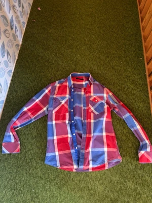 Rutig skjorta från Jack & Jones - Säljer en snygg rutig skjorta från Jack & Jones i rött, blått och vitt. Skjortan har långa ärmar, klassisk krage och knäppning framtill. Perfekt för dig som gillar en avslappnad men ändå stilren look.