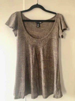 Glittrig H&M Topp m. Volangärm / Y2K Look – Stl XS - Fin glittrig topp från H&M i en varm brun/taupe ton med skimmer av guldtråd. Toppen har charmiga volangärmar, en djupare rund ringning och ett hålmönstrat parti upptill. Modellen är lite kortare baktill vilket ger en fin silhuett. Innehåller 11% ull som ger extra kvalitet. Snygg Y2K och boho-känsla som passar perfekt till både vardag och fest. Storlek: XS Mått: Ca 62 cm lång från axeln (lite kortare baktill) och axelbredd ca 33 cm Material: 63% akryl, 16% polyamid, 11% ull, 10% metallfibrer.