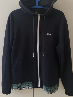 Svart hoodie med dragkedja från BOSS - Snygg svart hoodie från BOSS med dragkedja framtill och huva med dragsko. Diskret BOSS-logga på bröstet och upprepad logga längs nederkanten. Mjukt material och två fickor på sidorna. Perfekt för en chill och stilren look.