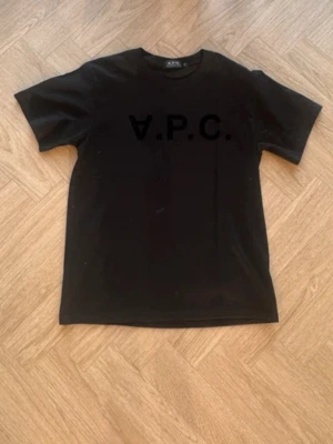 Svart t-shirt från A.P.C. - Snygg svart t-shirt från A.P.C. med klassisk rund hals och tryckt logga framtill. Tillverkad i mjuk bomull som känns skön mot huden. Perfekt till jeans eller shorts för en clean och enkel stil.