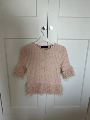 Ljusrosa kofta med fransdetaljer - Supersöt ljusrosa kofta med ribbad struktur, knappar framtill och unika fransdetaljer vid ärmslut och nederkant. Kortare modell med halvlånga ärmar, perfekt för att lyfta din outfit. Materialet känns mjukt och bekvämt.