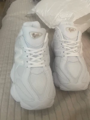 Vita New Balance 9060 sneakers strl 41 - Säljer ett par fräscha vita New Balance 9060 sneakers med chunky sula och klassisk N-logga på sidan. Skorna har en kombination av mesh och syntetmaterial för en sportig look och bra andningsförmåga. Perfekta för dig som gillar stilrena och bekväma sneakers.
