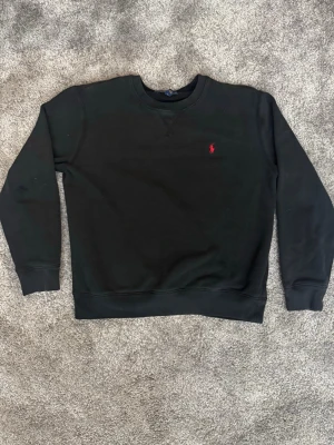 Svart sweatshirt från Polo Ralph Lauren - Klassisk svart sweatshirt från Polo Ralph Lauren med rund halsringning och den ikoniska röda logotypen broderad på bröstet. Observera att den är välanvänd och där med har ett litet märke på höger armbåge 