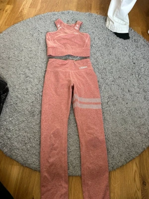 Rosa träningsset från Stronger - Säljer ett snyggt rosa träningsset från Stronger i storlek XS. Setet består av en sport-bh med breda axelband och matchande tights med vita detaljer på ena benet. Materialet är mjukt och stretchigt, 87% polyester och 13% elastan, perfekt för gym eller yoga.