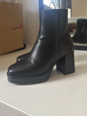 Svarta platåstövletter med klack - Säljer ett par svarta stövletter i slätt skinnimitation med chunky platå och bred hög klack. Modellen har rundad tå och dragkedja på insidan för enkel påtagning. Perfekta för dig som gillar en edgy och trendig look.
