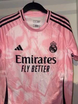 Rosa Real Madrid matchtröja Adidas - Säljer en rosa Real Madrid matchtröja från Adidas med svarta detaljer och klubbmärke på bröstet. Tröjan har korta ärmar, rund hals och är tillverkad i ett lätt, ventilerande material. Tryck med 'Emirates Fly Better' på framsidan och klassiska Adidas-ränder på axlarna.