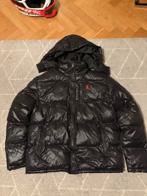 Svart dunjacka med huva från Polo Ralph Lauren - Snygg svart dunjacka från Polo Ralph Lauren med huva och dragkedja framtill. Jackan har två sidofickor med dragkedja och en bröstficka med röd logga. Perfekt för kalla dagar och riktigt skön att bära. Storlek L. 