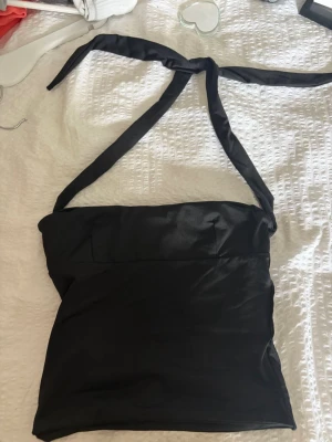 Svart halterneck topp i satin - Snygg svart halterneck topp i glansigt satinmaterial. Toppen har knytband i nacken och är ärmlös, vilket ger en stilren och trendig look. Perfekt att styla med jeans eller kjol för en cool outfit.
