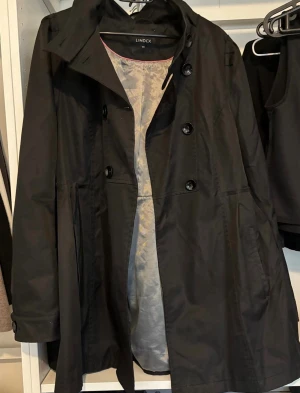 Svart trenchcoat från Lindex - Stilren svart trenchcoat från Lindex med dubbelknäppning och klassisk krage. Jackan har fickor på sidorna och ett lätt utställt snitt som ger en snygg siluett. Perfekt för dig som vill ha en tidlös och trendig look. Materialet är slätt och vattenavvisande.