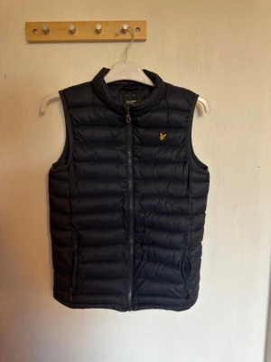 Lyle & Scott väst för barn - Snygg svart dunväst med quiltad design och ståkrage. Västen har dragkedja framtill, två fickor med dragkedja och en gul broderad logga på bröstet. Perfekt för lager-på-lager och håller dig varm under kyliga dagar.