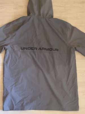 Grå vindjacka från Under Armour - Snygg grå vindjacka från Under Armour med huva och stort logotryck på ryggen. Jackan har långa ärmar och är tillverkad i ett lätt, vindtätt material som passar perfekt för blåsiga dagar. (Obs den här en stor olja fläck där back, samt några framme) men dom är inte så märkbara speciellt på kvällarna.