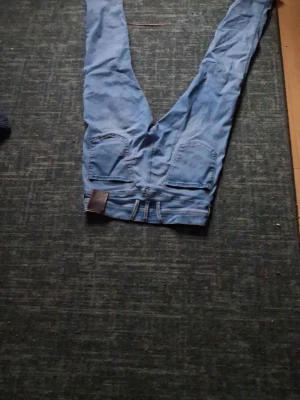 Blå raka jeans med fem fickor - Säljer ett par klassiska blå jeans med rak passform och fem fickor. Jeansen har bälteshällor och en läderpatch bak i midjan. Tillverkade i denim med en snygg tvättad look. Perfekta för en avslappnad stil.