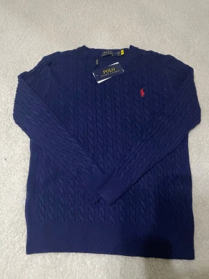 Mörkblå kabelstickad tröja Polo Ralph Lauren - Snygg mörkblå kabelstickad tröja från Polo Ralph Lauren med rund halsringning och den klassiska röda logotypen broderad på bröstet. Tröjan har långa ärmar och ribbade muddar vid ärmslut och nederkant. Perfekt för en stilren look. Storleken är S men den passats också XS