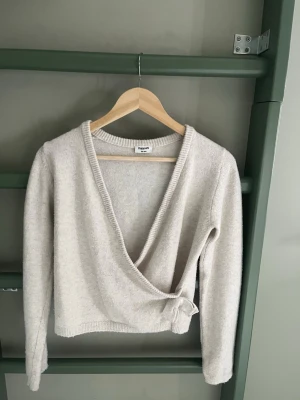 Beige omlottkofta från Kappahl - Mysig beige stickad omlottkofta från Kappahl med knytband i sidan. Koftan har lång ärm och ribbad kant vid halsringningen. Perfekt för lager på lager och enkel att matcha med olika outfits.