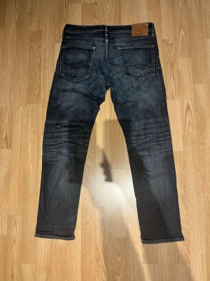 Mörkblå jeans från Jack & Jones - Snygga mörkblå jeans från Jack & Jones med klassisk femficksdesign och raka ben. Jeansen har kontrastsömmar och en läderpatch bak i midjan. Materialet är denim med lätt tvättad look och subtila slitningar för en trendig känsla. Finns ett litet lagat hål på knät