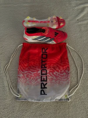 Adidas Predator Elite röda fotbollsskor och påse till - Säljer ett par grymma Adidas Predator elite fotbollsskor i röd med svarta och silvriga detaljer och även en påse till. Skorna har låg profil, snörning och dobbar för spel på gräs. Medföljer en matchande röd/vit påse med Predator-tryck. Perfekta för dig som vill sticka ut på planen!