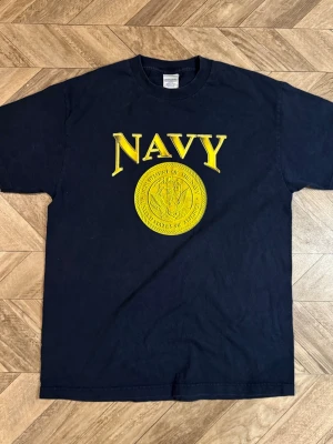 Marinblå t-shirt med gul NAVY-tryck - Säljer en marinblå t-shirt med stort gult NAVY-tryck och emblem på bröstet. Klassisk passform och rund halsringning. Perfekt för dig som gillar enkel och stilren design med militärvibbar.