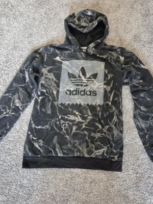 Svart och grå hoodie från Adidas - Säljer en svart och grå hoodie från Adidas med ett stort tryck av Adidas-loggan på bröstet. Hoodien har ett marmorerat mönster, huva och en stor magficka. Muddar vid ärmslut och nederkant. Perfekt för en avslappnad streetwear-look.