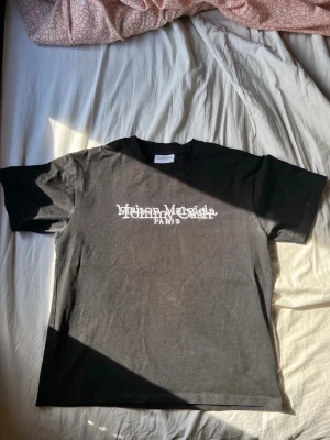 Svart Maison Margiela t-shirt - Säljer en svart t-shirt från Maison Margiela med vit logga och texten 'PARIS' tryckt på bröstet. Klassisk passform och rund halsringning. Perfekt för dig som gillar stilrena och trendiga plagg.