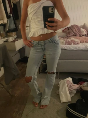 Ljusblå slitna bootcut jeans - Säljer ett par ljusblå jeans med bootcut passform och slitningar vid knäna. Jeansen har klassisk femficksdesign och är tillverkade i denim med en lätt tvättad look. Perfekta för en avslappnad och trendig stil. Väldigt slitna längst ner vid hälen men inget som märks när de är på