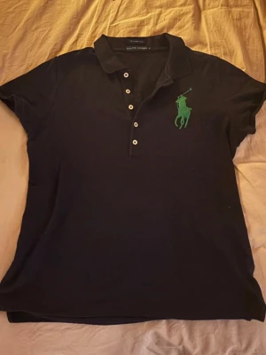 Svart pikétröja från Ralph Lauren - Klassisk svart pikétröja från Ralph Lauren med grönt broderat logotyp på bröstet. Tröjan har korta ärmar, krage och knappar framtill. Tillverkad i mjuk bomull som känns skön mot huden. Perfekt för en avslappnad men ändå stilren look.