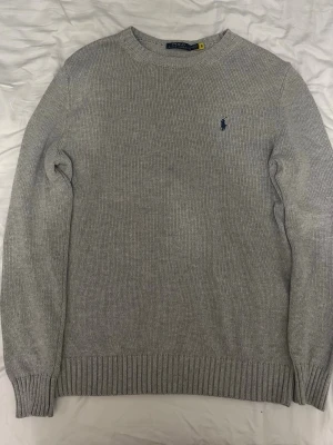 Grå stickad tröja från Polo Ralph Lauren - Klassisk grå stickad tröja från Polo Ralph Lauren med rund halsringning och diskret blå logga broderad på bröstet. Tröjan har långa ärmar och ribbade muddar vid ärmslut och nederkant. Perfekt basplagg till garderoben.