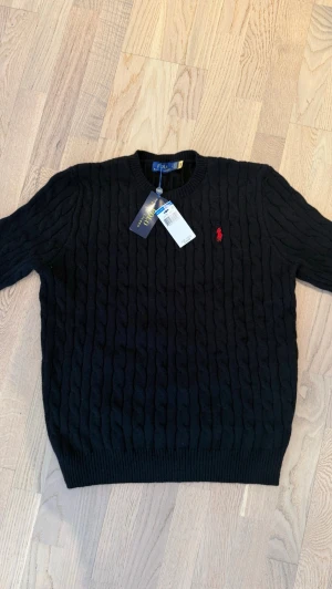 Svart kabelstickad tröja Polo Ralph Lauren - Svart kabelstickad tröja från Polo Ralph Lauren med rund halsringning och röd broderad logga på bröstet. Tröjan har ribbstickade muddar vid ärmslut och nederkant. Klassisk och tidlös design i mjukt material.
