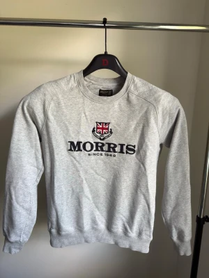 Grå sweatshirt från Morris - Snygg grå sweatshirt från Morris med broderad logga och brittisk flagga på bröstet. Tröjan har rund halsringning, ribbade muddar vid ärmslut och nederkant samt en klassisk passform. Perfekt för en avslappnad och stilren look.                                                 Varan är använd och har två mindre defekter: en liten, ljus smutsfläck ovanför "O":et i loggan, samt ett litet hål i tyget strax ovanför fläcken (se närbild). I övrigt i normalt använt skick. Säljs därför till reducerat pris. 