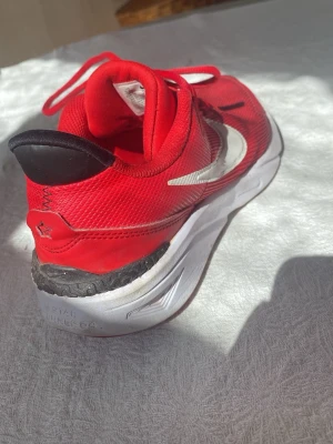 Röda Nike sneakers med vit sula - Snygga röda Nike sneakers med vit sula och svart detalj på hälen. Skon har en vit swoosh-logga på sidan och är tillverkad i meshmaterial för bra andningsförmåga. Perfekta för dig som vill ha en sportig och trendig look.