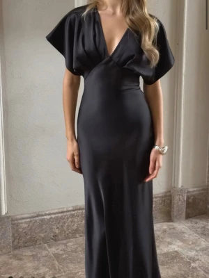 Svart långklänning från Gina Tricot - Elegant svart långklänning från Gina Tricot med djup v-ringning och vida, korta ärmar. Klänningen har en figurnära passform och är tillverkad i ett glansigt, mjukt material som ger en lyxig känsla. Perfekt för dig som vill sticka ut med en stilren look.